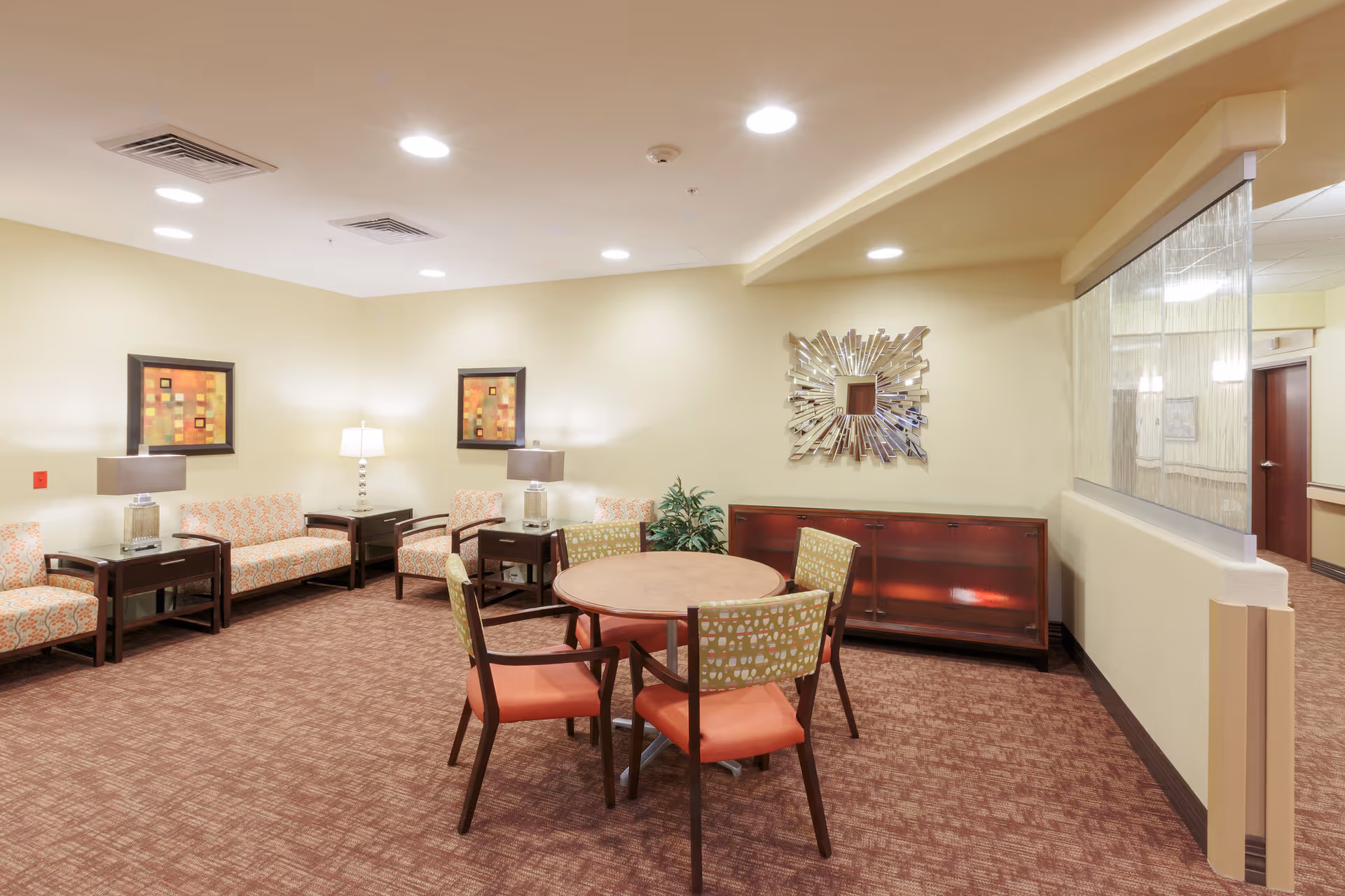A well-lit common area with a round table surrounded by four chairs with orange cushions and patterned backs. Along the walls are several cushioned armchairs and sofas with patterned upholstery, dark wooden side tables with lamps, two framed abstract artworks, a decorative mirror, and a wooden cabinet. The room has beige walls and a brown carpeted floor.