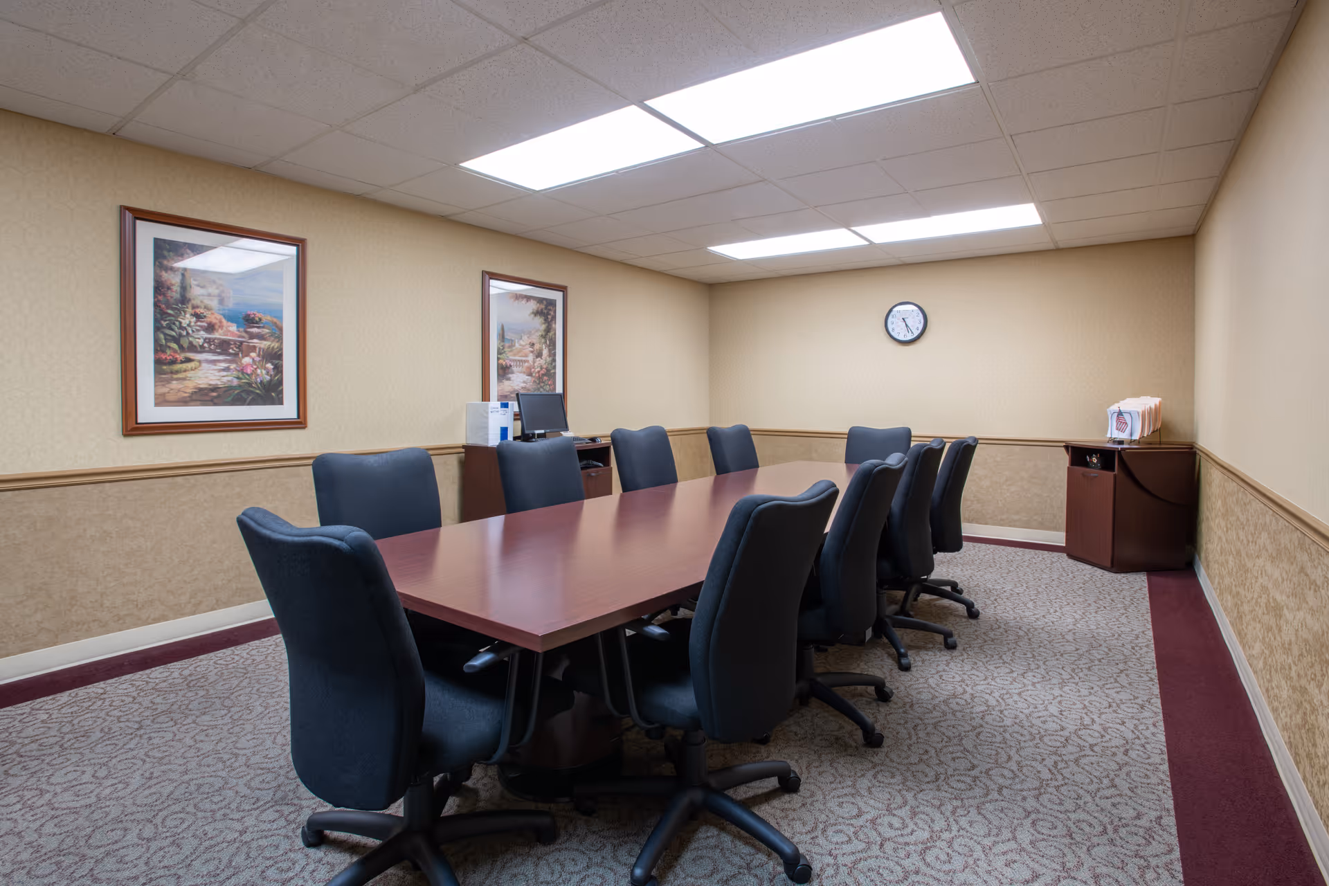A conference room with a long rectangular wooden table surrounded by ten black office chairs on wheels. The room has beige walls with two framed landscape paintings, a wall clock, and a small cabinet with files and a computer monitor on top. The ceiling has fluorescent lighting panels.