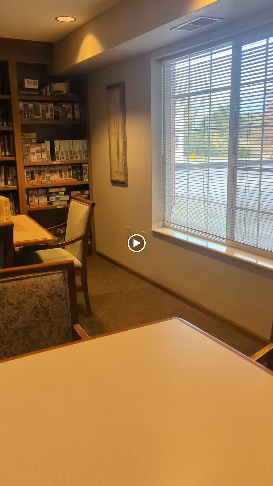 Interior view of a room with a table and chairs, a bookshelf filled with board games, a framed picture on the wall, and a large window with blinds letting in natural light.
