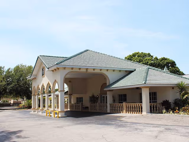 Exterior view of a single-story building with a green tiled roof and multiple arches along the front. The building has a covered entrance area with columns and a paved driveway in front. Trees and shrubs surround the building under a clear sky.