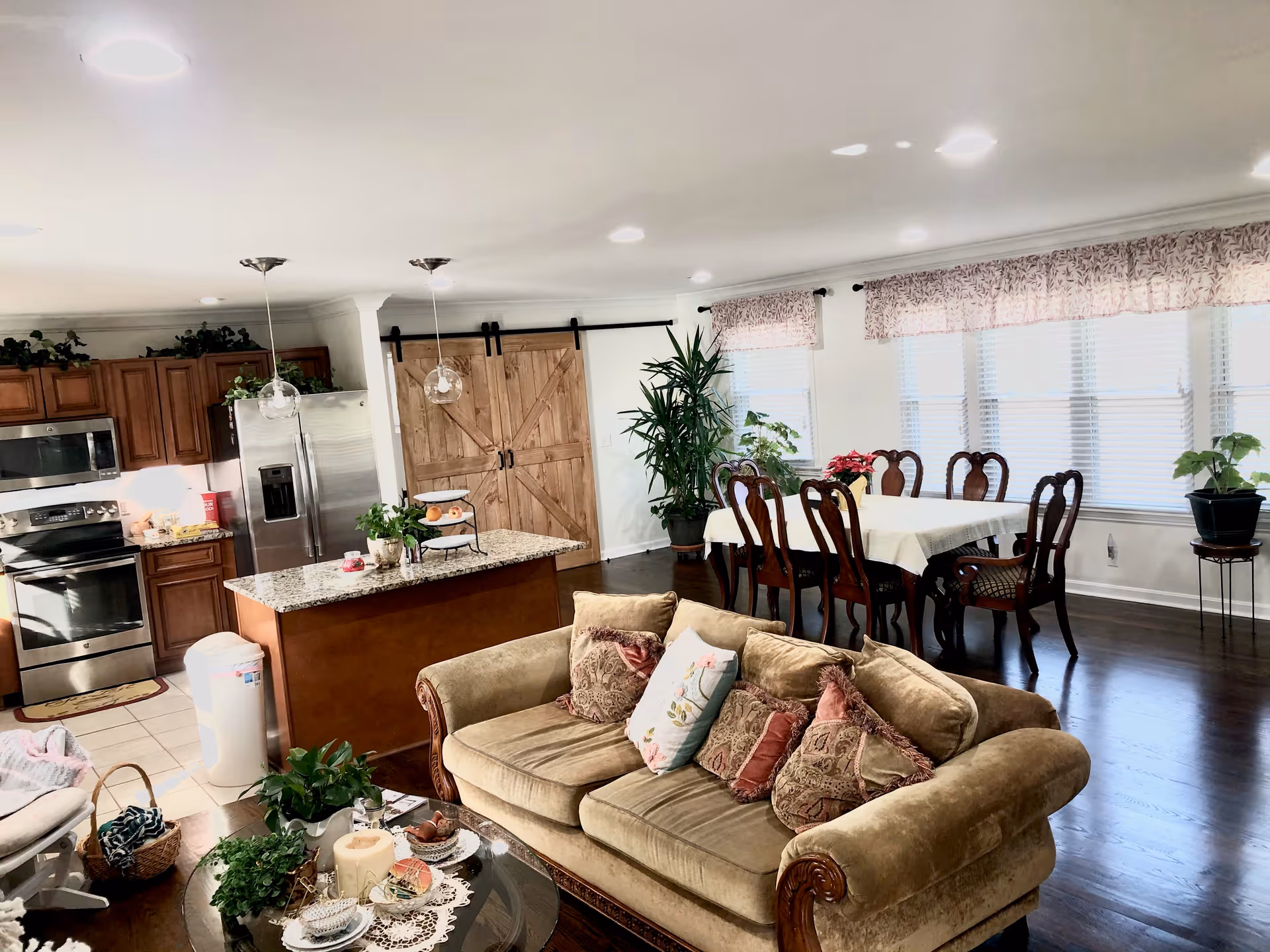 A bright and spacious open-concept living area featuring a beige sofa with multiple decorative pillows, a glass coffee table with plants and candles, a wooden dining table with six chairs covered with a white tablecloth, and a kitchen area with wooden cabinets, stainless steel appliances, and a granite island. Large windows with patterned valances allow natural light to fill the room, and there are several potted plants throughout the space. A rustic wooden sliding barn door is visible near the kitchen.