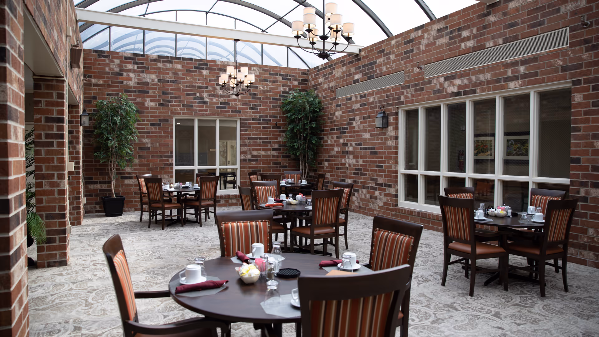 Indoor dining area with round wooden tables and striped cushioned chairs set with cups, glasses, and napkins. The room has brick walls, large windows, two tall potted plants, and a glass ceiling with chandeliers providing natural light.