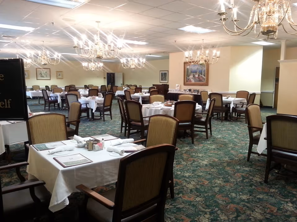 A spacious dining room with multiple tables covered in white tablecloths, each set with napkins, menus, and condiments. The room features green patterned carpet, beige walls adorned with framed paintings, and several elegant chandeliers hanging from the ceiling. Wooden chairs with cushioned seats surround the tables.