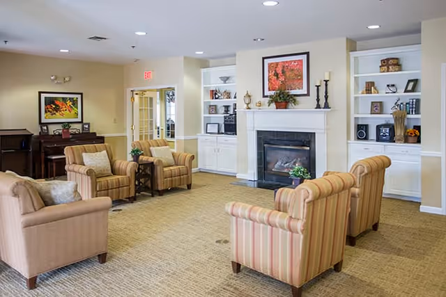 A cozy living room area in a senior living facility with four striped armchairs and one solid armchair arranged around a fireplace. The fireplace has a framed floral picture above it and built-in shelves on either side holding decorative items. There is a piano with a framed picture on the wall behind it, and a door leading to another room in the background.