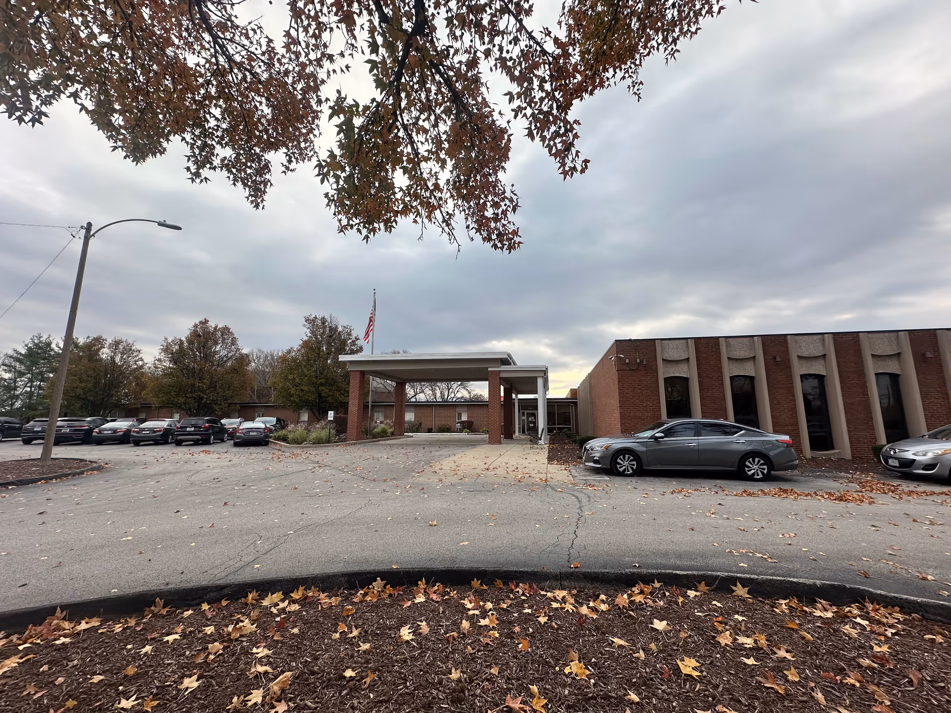 Exterior view of a single-story brick building with a covered entrance and an American flag on a flagpole. Several cars are parked in the parking lot in front of the building. Trees with autumn leaves frame the top of the image and fallen leaves cover the ground in the foreground.