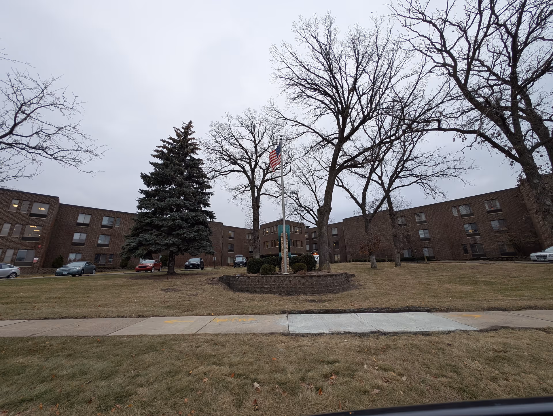 Front view of a low-rise brick senior care building with an American flag, leafless trees, parked cars, and a grassy lawn under an overcast sky.