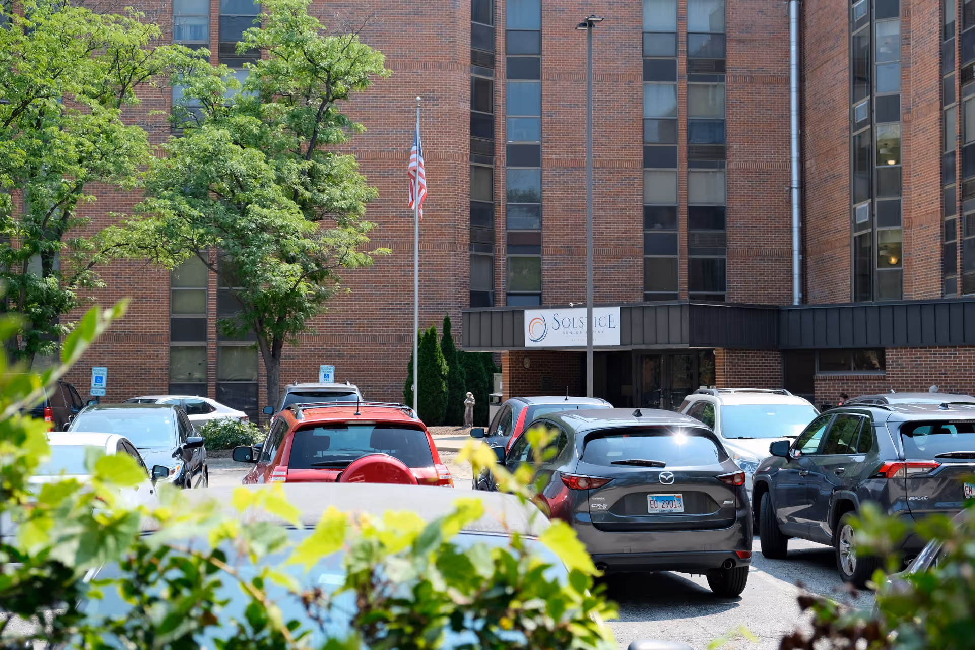 View of the exterior entrance of Solstice Senior Living at Palatine, showing a brick building with multiple windows, a parking lot with several cars, green trees, and an American flag on a flagpole near the entrance.