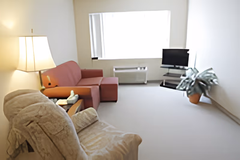 A living room with a beige recliner chair covered with a light fabric, a wooden side table with a lamp and decorative items, a red sofa with an ottoman, a flat-screen TV on a stand, a potted plant, and a large window with vertical blinds letting in natural light.