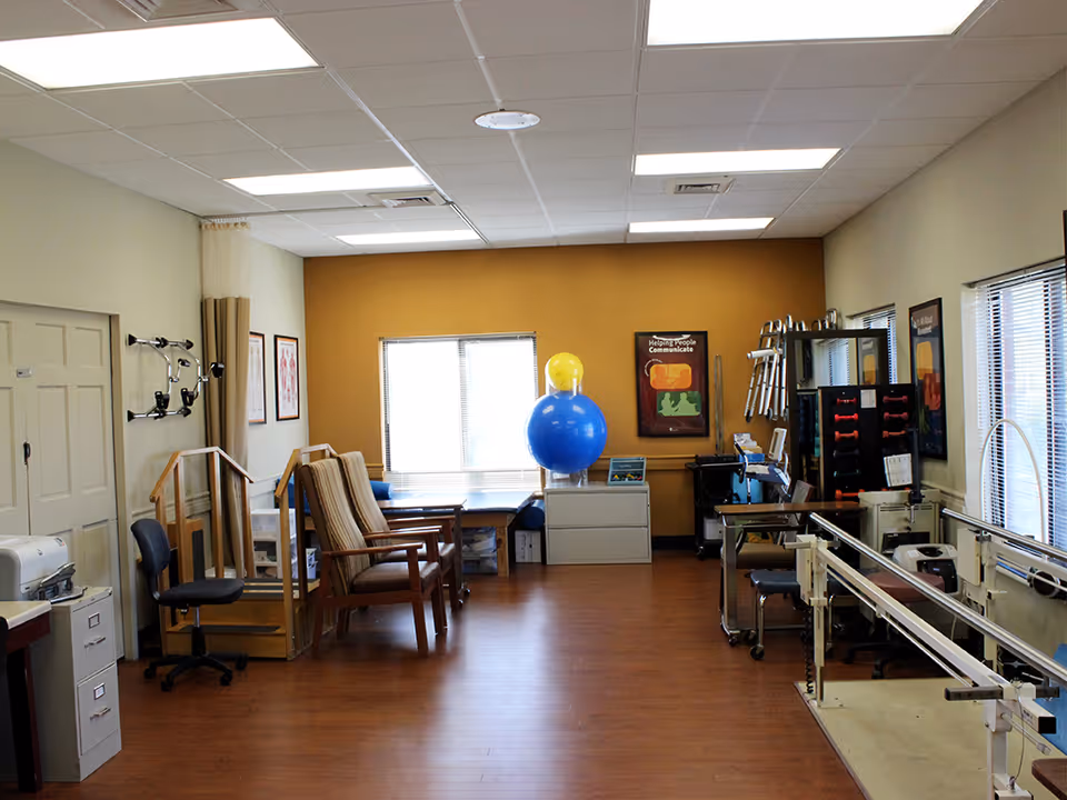 A therapy or rehabilitation room with wooden flooring, beige and yellow walls, and fluorescent ceiling lights. The room contains exercise equipment including parallel bars, therapy balls, chairs, and various physical therapy tools. There are windows with blinds on the far wall and the right side, and framed posters on the walls.