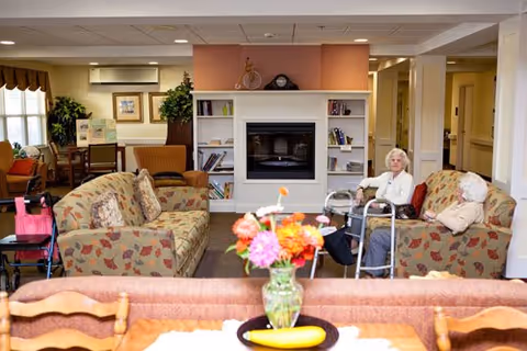 A cozy living room area in a senior living facility with two elderly women sitting on floral patterned sofas near a fireplace with built-in bookshelves. There is a vase with colorful flowers on a table in the foreground, and the room is warmly lit with various chairs and plants in the background.
