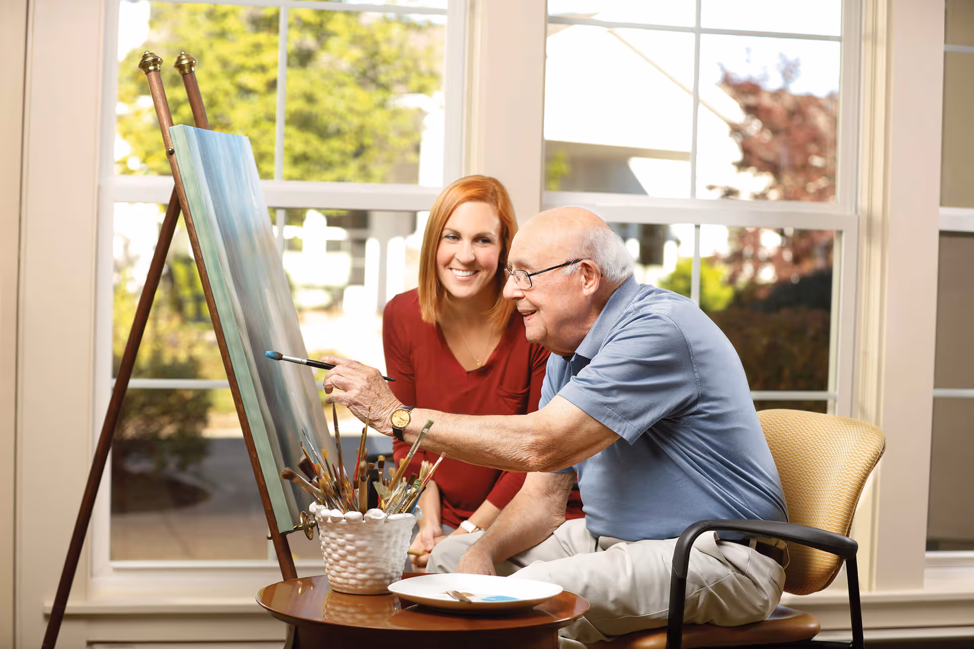 An elderly man painting on a canvas set on an easel while a younger woman sits beside him smiling, both in a bright room with large windows showing greenery outside.