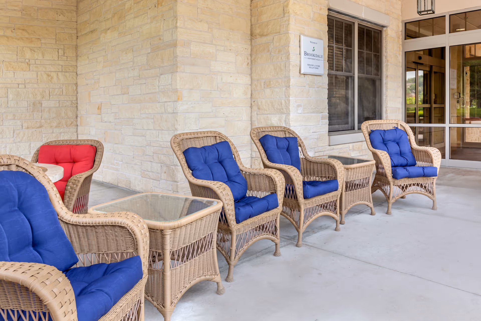 Outdoor seating area at Brookdale Northwest Hills featuring wicker chairs with blue and red cushions arranged along a stone wall near a window and entrance door.
