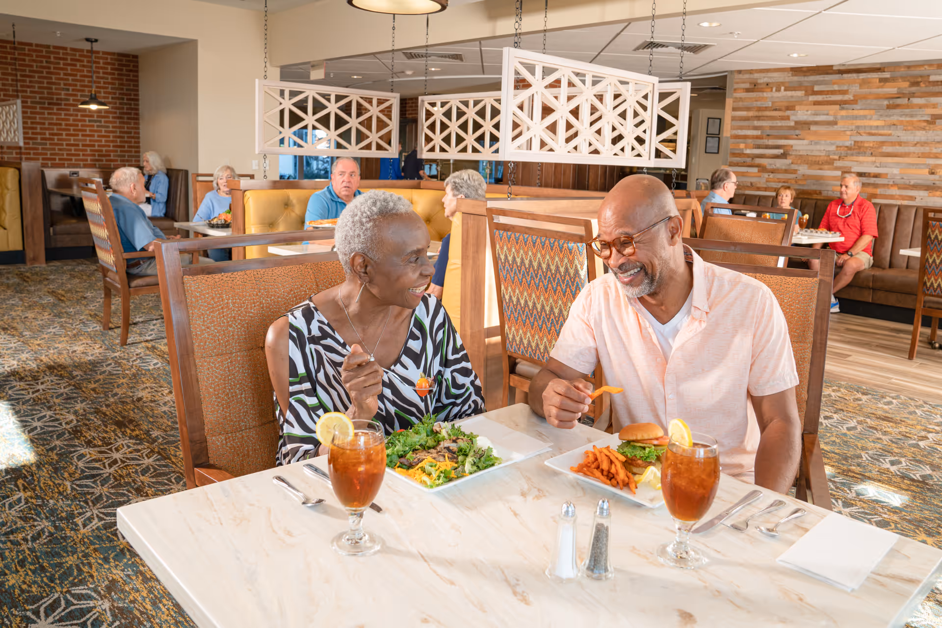 Two elderly people, a woman and a man, are sitting at a table in a dining room enjoying a meal together. The woman is eating a salad while the man is eating a burger with sweet potato fries. Both have glasses of iced tea with lemon. Other elderly people are seated at tables in the background, and the room has warm lighting, patterned carpet, and wooden furniture with cushioned seats.