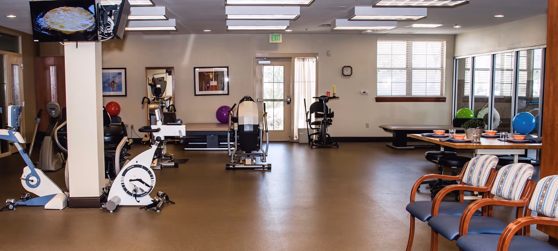 A spacious room with exercise equipment including stationary bikes and weight machines, a table set with dishes and chairs, and several colorful exercise balls. The room has large windows, a door with a window, and bright overhead lighting.