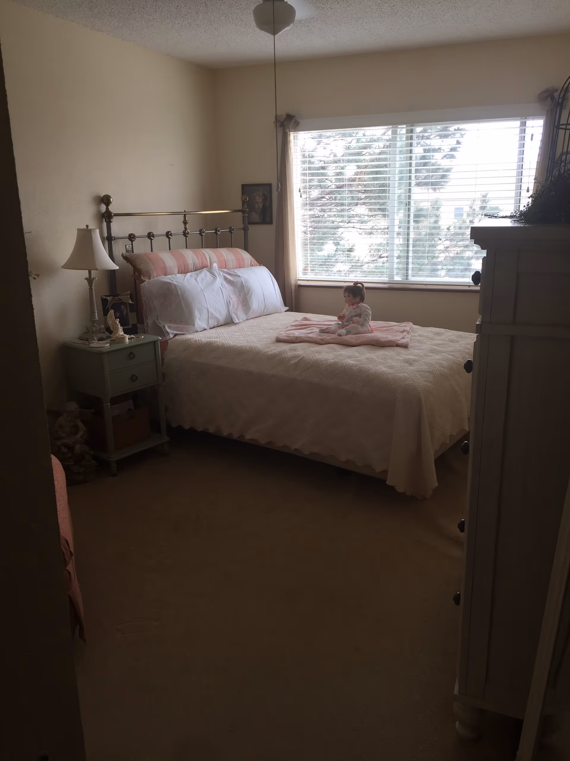 A cozy bedroom with a bed covered in a light-colored quilt and pillows, a small doll sitting on a folded blanket on the bed. There is a window with blinds letting in natural light, a nightstand with a lamp and decorative items, and a white dresser on the right side of the room.