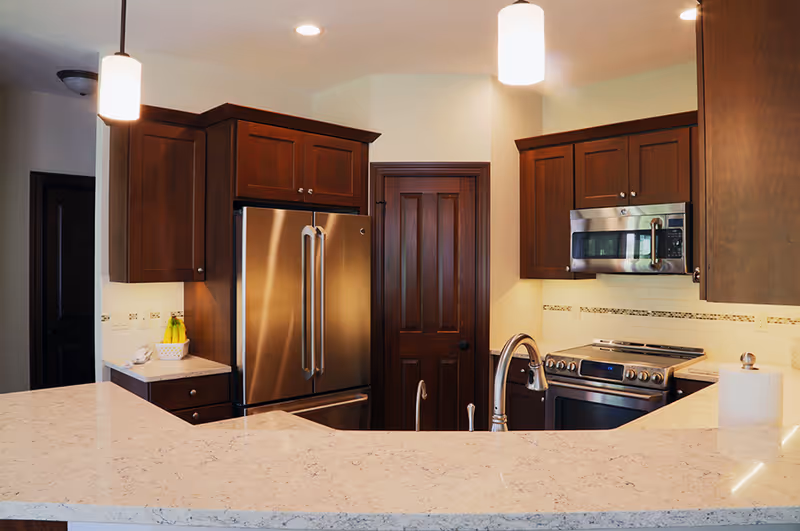 Modern kitchen with dark wood cabinets, stainless steel refrigerator, microwave, and stove. The kitchen features a light-colored marble countertop with a sink and a paper towel holder. Two pendant lights hang from the ceiling.