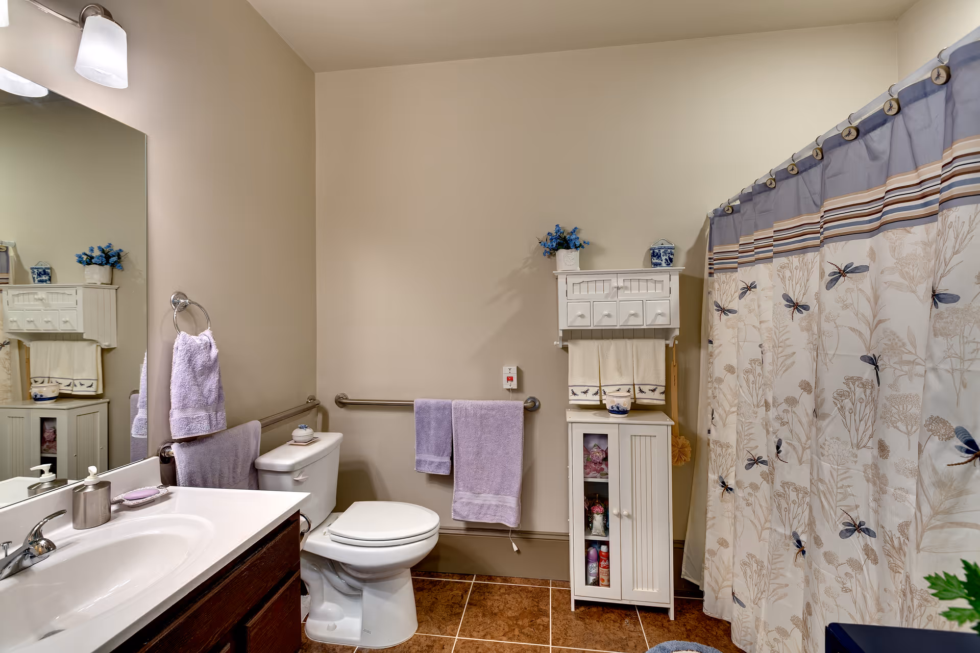 A bathroom with a white toilet, a sink with a countertop and faucet, a large mirror above the sink, and a shower with a curtain featuring a dragonfly and floral pattern. There are purple towels hanging on a towel ring and a grab bar, a white cabinet with decorative items and towels, and a tiled floor.