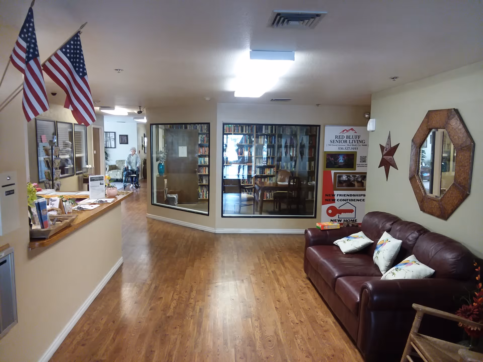 Interior view of a senior living facility hallway with wooden flooring, a reception desk on the left with two American flags, a leather couch with pillows on the right, a decorative mirror and star on the wall, and a glass-enclosed library area in the center background. An elderly person with a walker is visible in the distance.