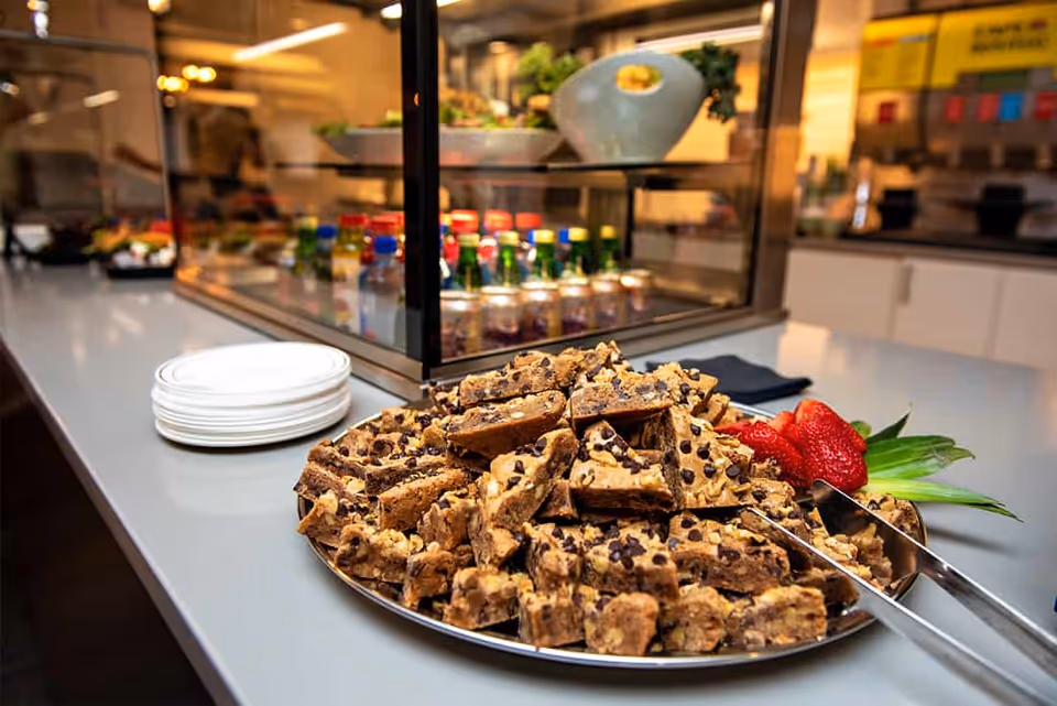 A plate piled with chocolate chip cookie bars and garnished with strawberries and pineapple leaves on a countertop in a dining area. Behind the plate, there are stacks of white plates and a glass display case with bottled beverages inside.