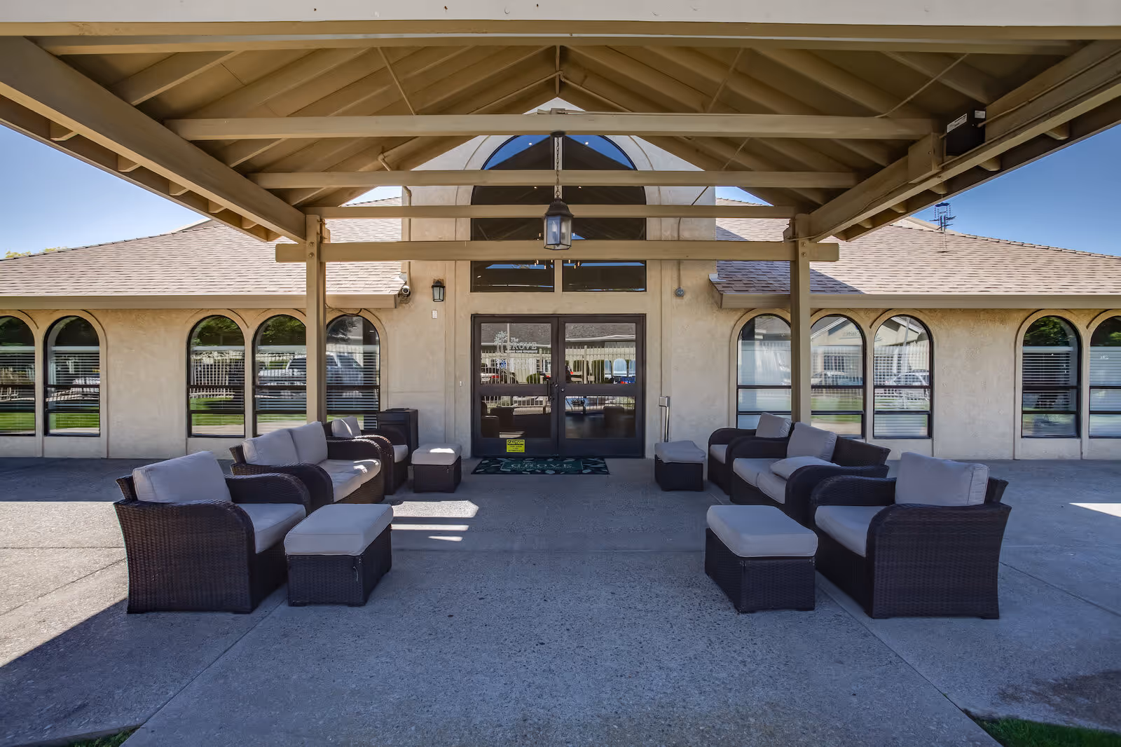 Covered front entrance of a senior living facility with wicker lounge seating arranged under a canopy in front of glass double doors.