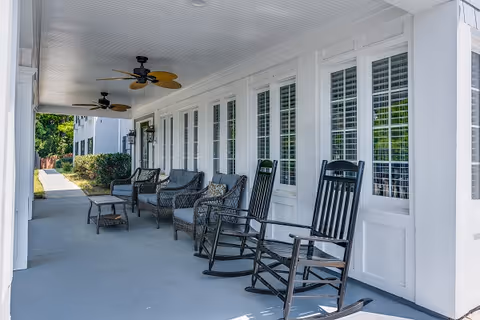 Covered outdoor porch area with several cushioned wicker chairs, two black rocking chairs, ceiling fans, and white-framed windows with shutters along the wall.