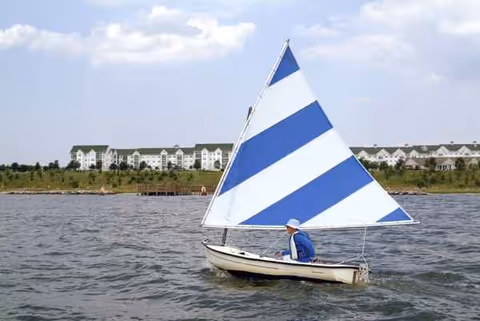 A small sailboat with a person gliding on calm water near waterfront buildings.