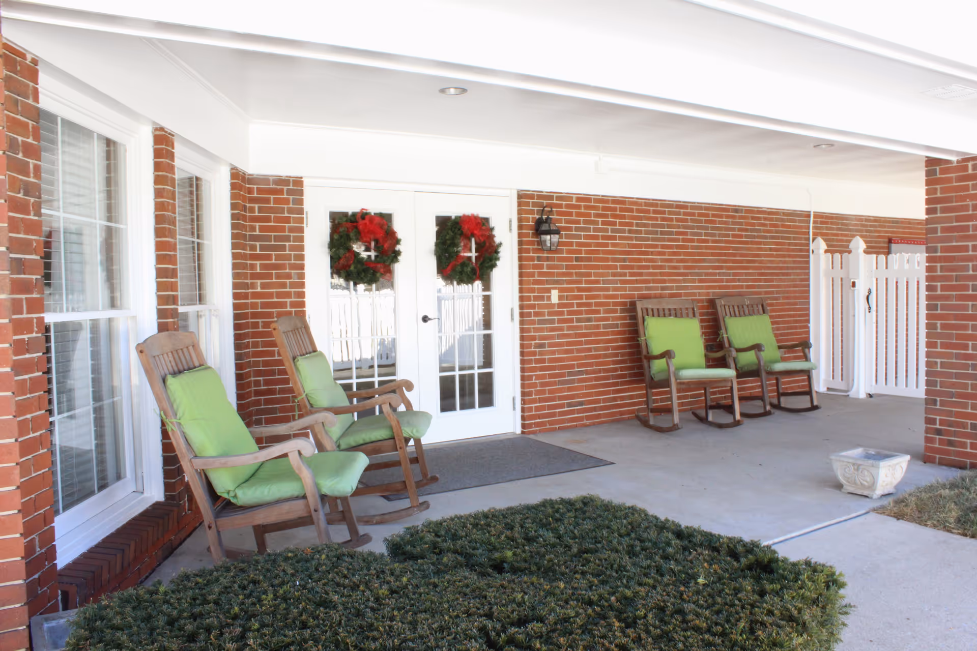 Covered outdoor patio area with four wooden rocking chairs with green cushions arranged near a brick wall and white double doors decorated with red and green wreaths. There is a white picket fence and some greenery in the foreground.