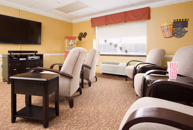 A cozy lounge area with beige upholstered armchairs arranged in two rows facing a large flat-screen TV mounted on a yellow wall. There is a dark wooden side table with a popcorn container on it, a popcorn machine in the corner, and movie-themed wall decorations near a window with a red valance and small potted plants on the windowsill.