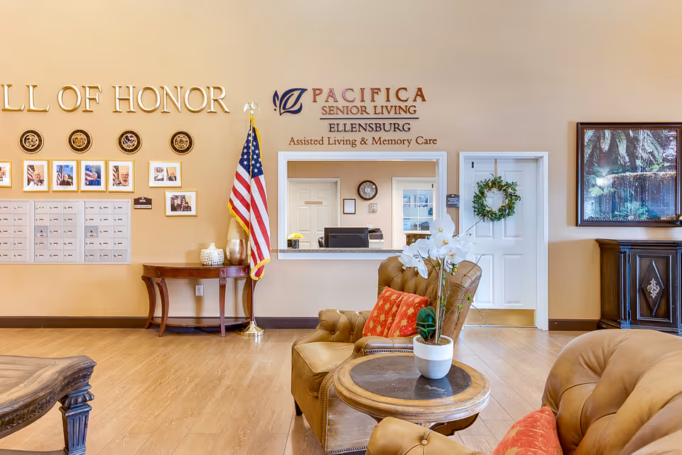 Interior view of a senior living facility lobby with comfortable leather chairs, a small round table with a white orchid plant, a wooden table with decorative items, an American flag, a mail area with multiple mailboxes, and a wall displaying a 'Wall of Honor' with photos and plaques. The wall also features the facility name 'Pacifica Senior Living Ellensburg Assisted Living & Memory Care'.