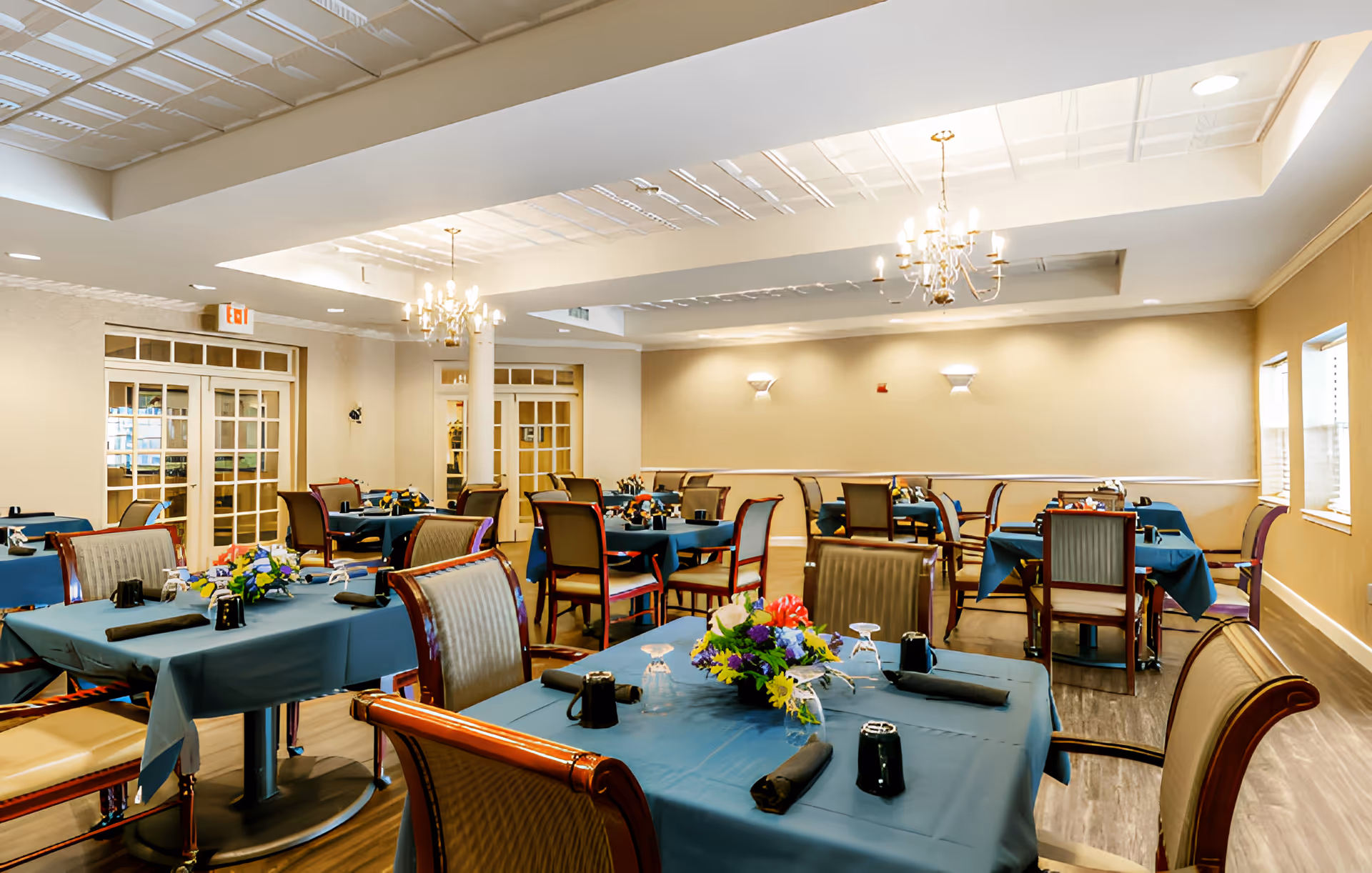 A bright and spacious dining room with multiple tables covered in blue tablecloths, each set with black napkins, glasses, and floral centerpieces. The room features wooden chairs with cushioned seats, chandeliers hanging from the ceiling, and large windows allowing natural light to enter.