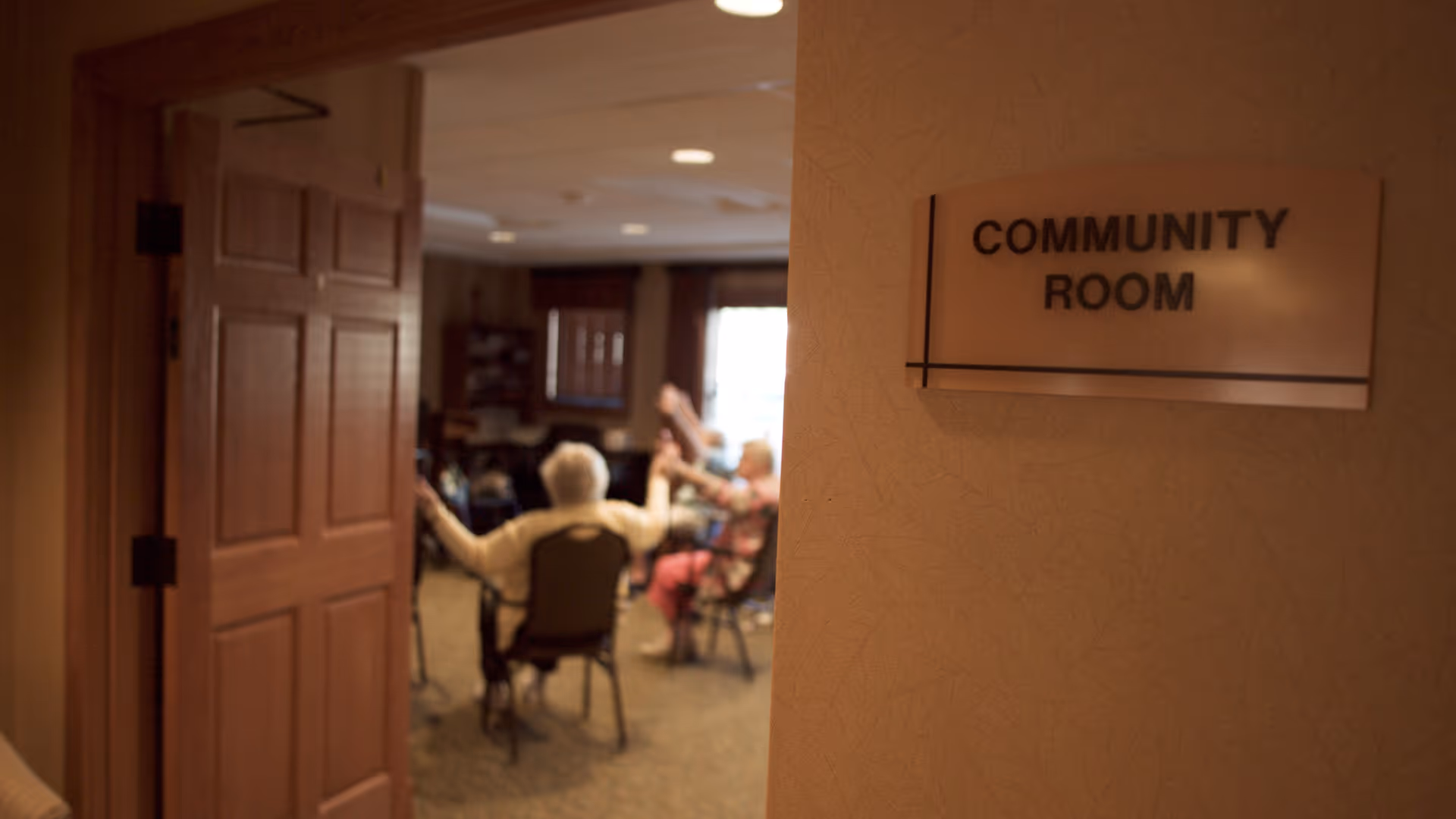 Open doorway shows a community room sign and a group of seated seniors participating in an activity.