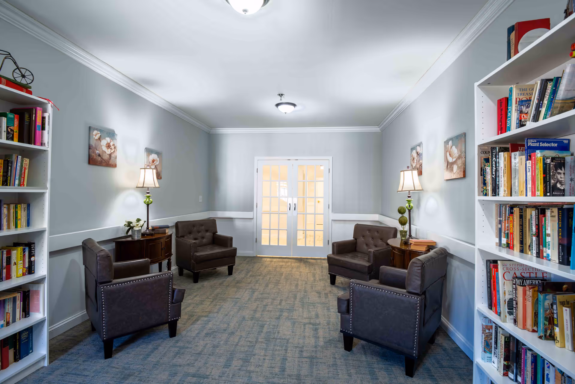 A cozy reading or sitting area in a senior living facility with four dark leather armchairs arranged in pairs facing each other. Each pair is separated by a small wooden side table with a lamp and decorative items. The room has light gray walls, two floral paintings, a blue carpet, and white double doors with glass panes at the far end. Bookshelves filled with books line both sides of the room.
