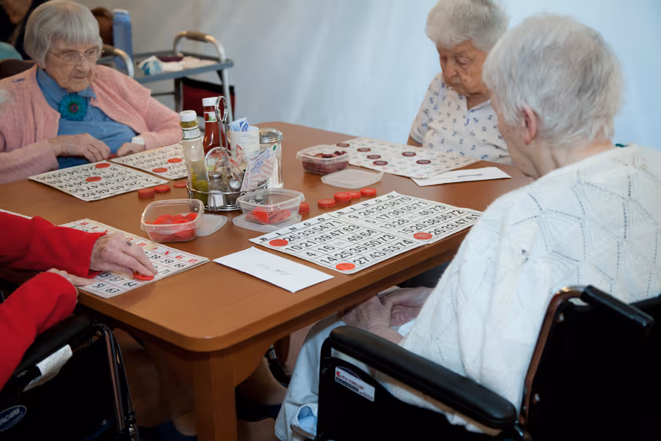 Four elderly women sitting around a wooden table playing bingo, with bingo cards and red markers in front of them. Two of the women are in wheelchairs, and there are condiments and containers in the center of the table.