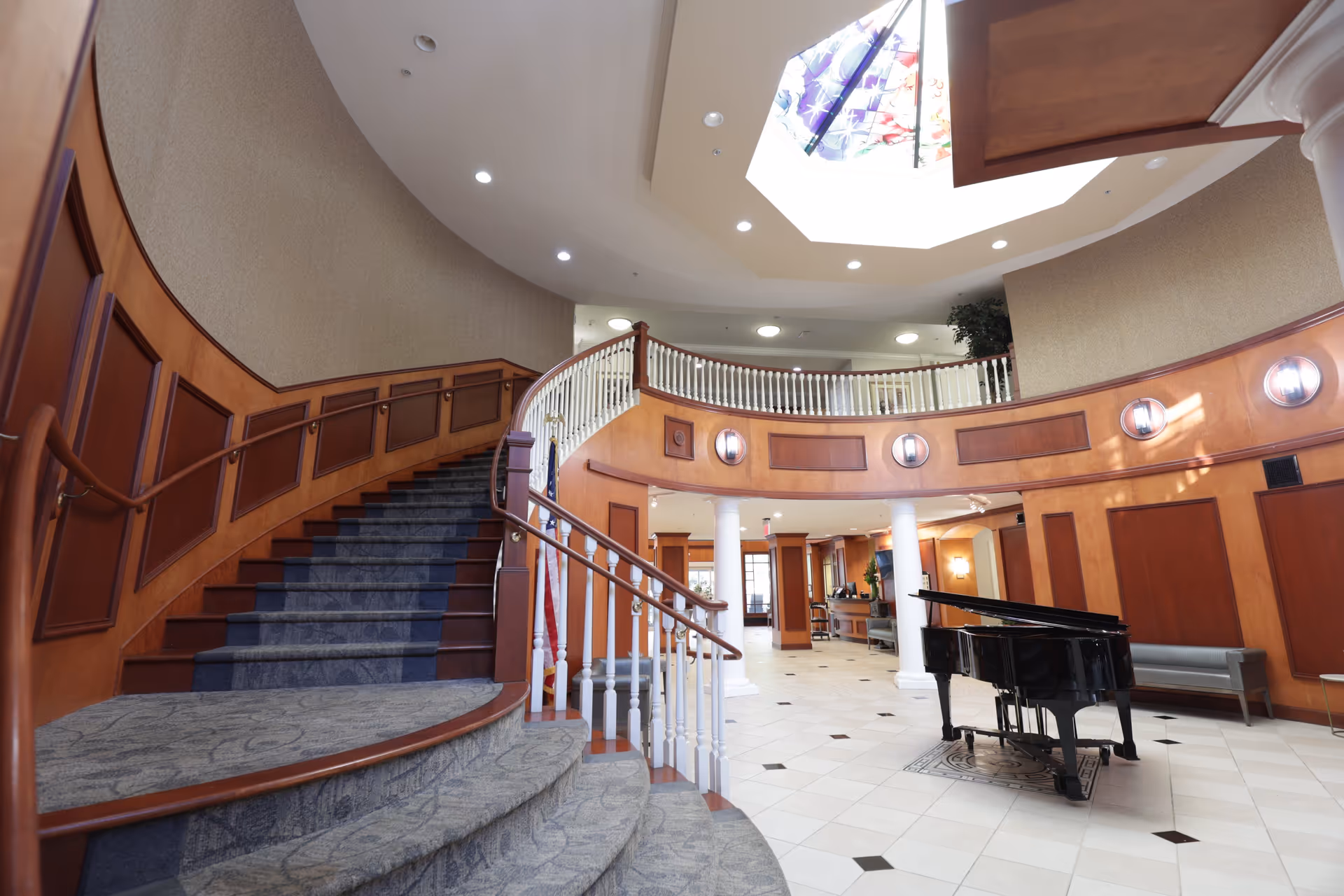 Interior view of a senior living facility lobby featuring a curved staircase with carpeted steps and wooden handrails, a black grand piano on a tiled floor, white columns, wood-paneled walls, and a skylight with stained glass above.