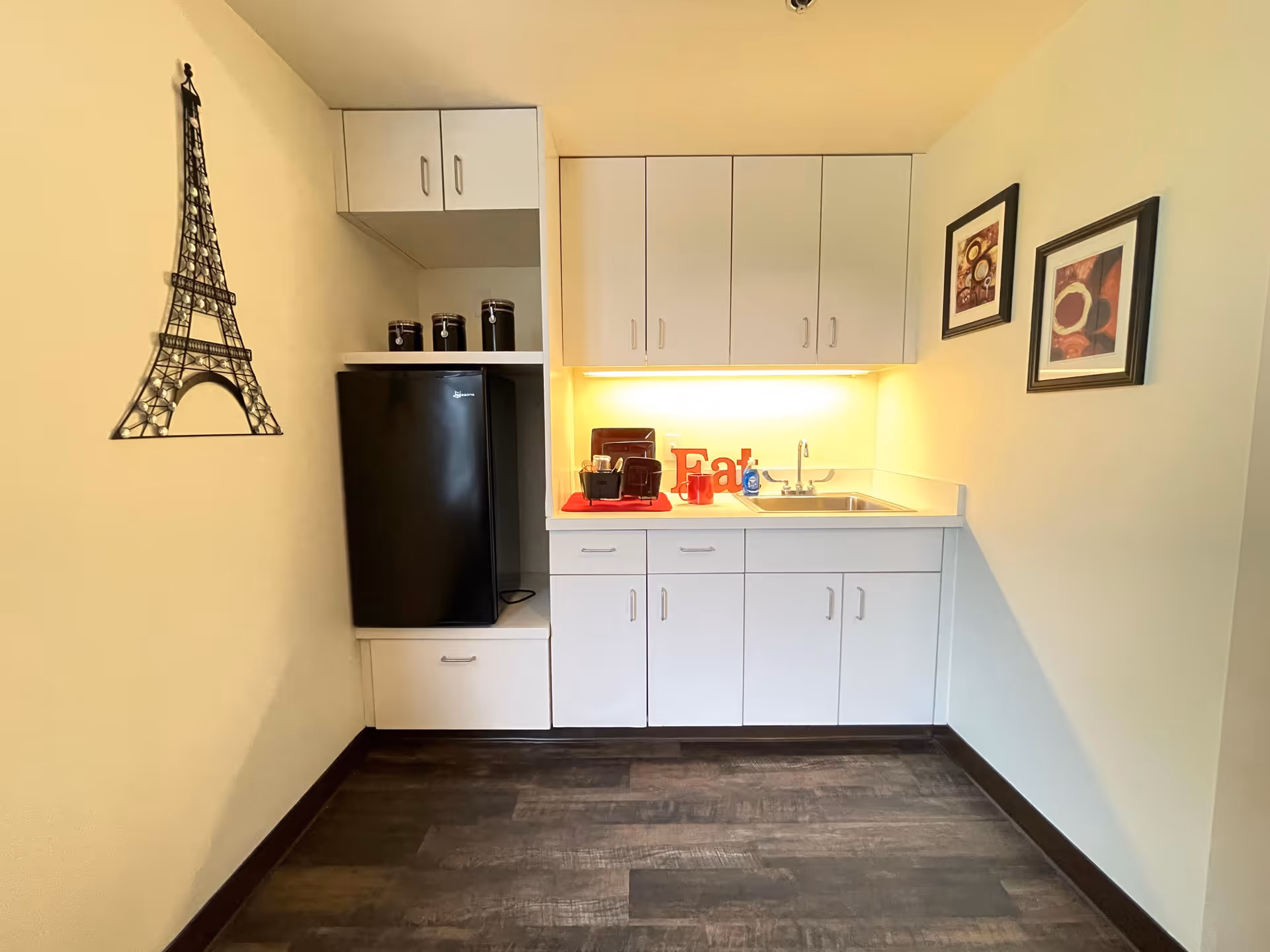 A small kitchenette area with white cabinets, a black mini refrigerator, a stainless steel sink, and a countertop with a red mat holding a coffee maker, mugs, and a decorative 'Eat' sign. The walls are light-colored, with two framed pictures on the right wall and a metal Eiffel Tower wall decoration on the left wall. The floor is dark wood.