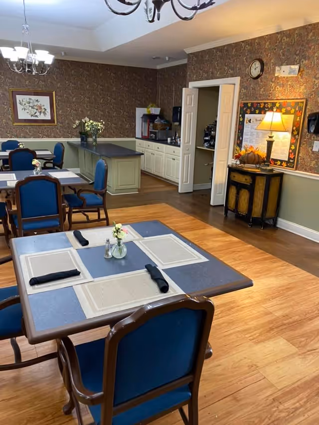 A dining room with several tables set with placemats, black napkins, and small flower vases. The room has wooden flooring, blue cushioned chairs, and a chandelier hanging from the ceiling. There is a counter area with cabinets and a coffee machine in the background, along with a small side table with a lamp and decorative items against a wallpapered wall.