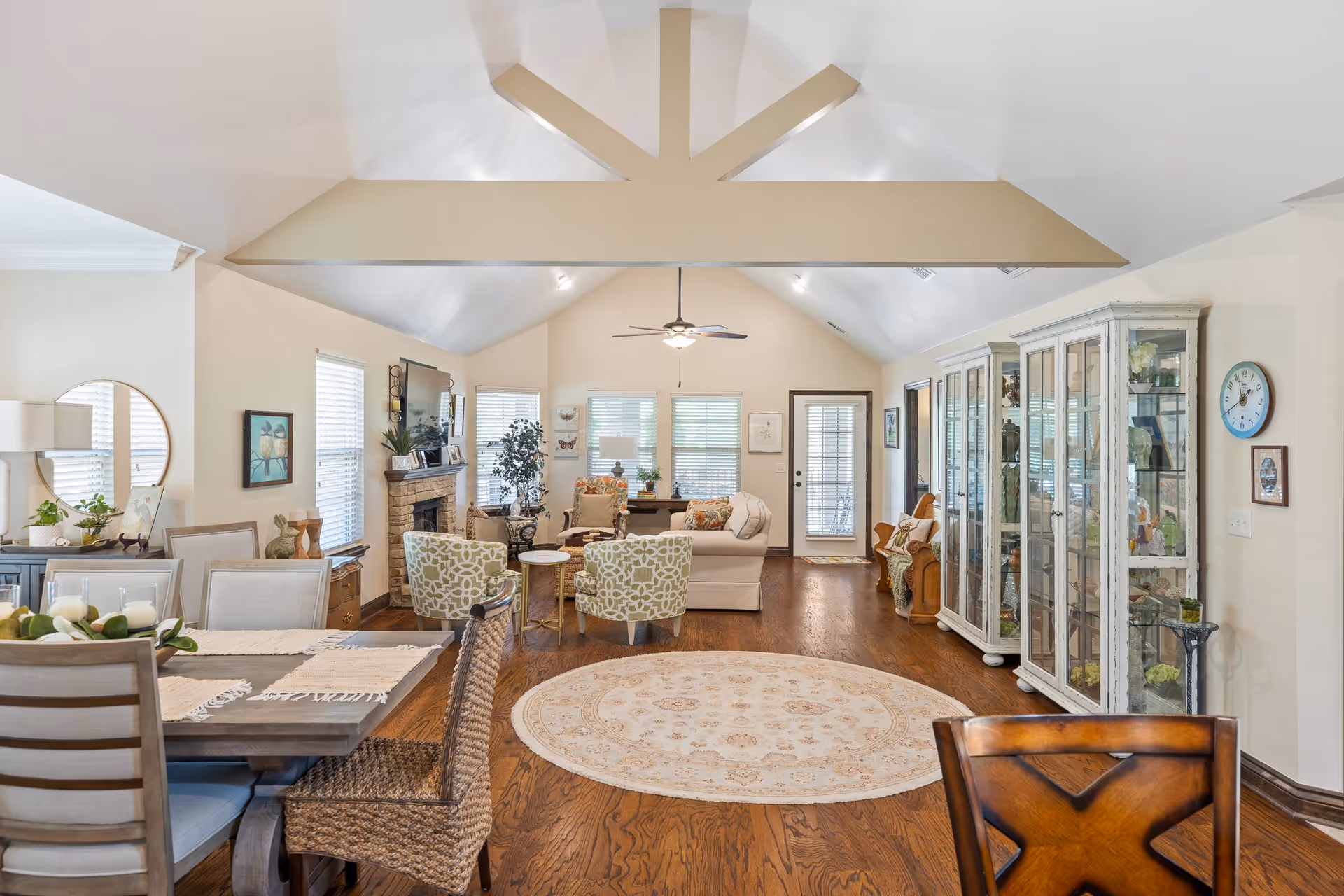 Open-concept living and dining area with a vaulted ceiling, seating around a fireplace, dining table, glass display cabinets, and hardwood floors.