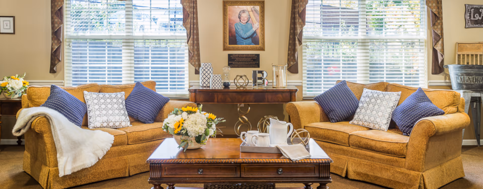 A cozy living room with two mustard yellow sofas adorned with blue and white patterned pillows. A wooden coffee table with a floral arrangement and a tea set sits in the center. Behind the sofas are large windows with white blinds and brown curtains. A wooden console table with decorative items and a framed portrait hangs on the wall between the windows.