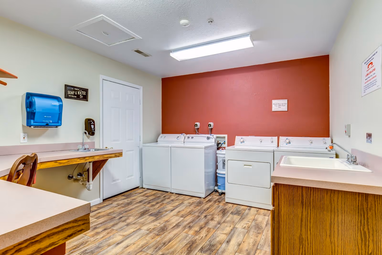 Laundry room with two washing machines and two dryers against a red accent wall. There is a countertop with a sink on the right side and a wooden chair next to a folding counter on the left. The floor has wood-patterned vinyl flooring and the walls are painted white and red. A blue paper towel dispenser and a soap dispenser are mounted on the left wall.