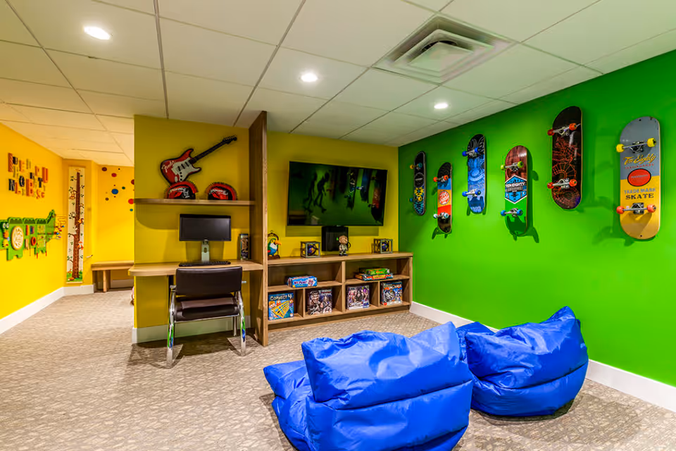 A colorful recreational room with a bright green wall displaying seven skateboards mounted vertically. In front of the wall are two blue bean bag chairs. A wooden entertainment unit holds board games and a flat-screen TV mounted above it. To the left, a small desk with a computer and chair is set against a yellow wall with a red guitar decoration above. The room has a carpeted floor and a drop ceiling with recessed lighting.