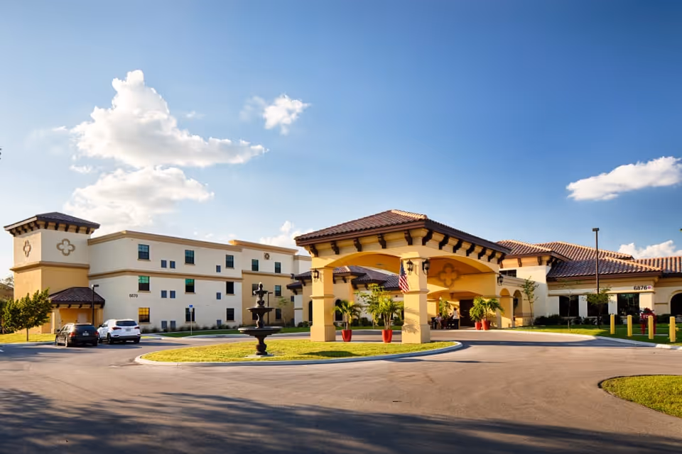 Exterior view of Discovery Commons Cypress Point facility featuring a large entrance with a covered driveway, a central fountain, and a three-story building with beige and yellow walls under a blue sky with scattered clouds.