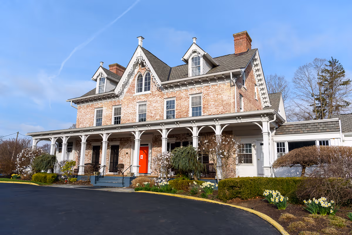 Historic two-story brick building with a covered wraparound porch, red front door, and landscaped driveway under a blue sky.
