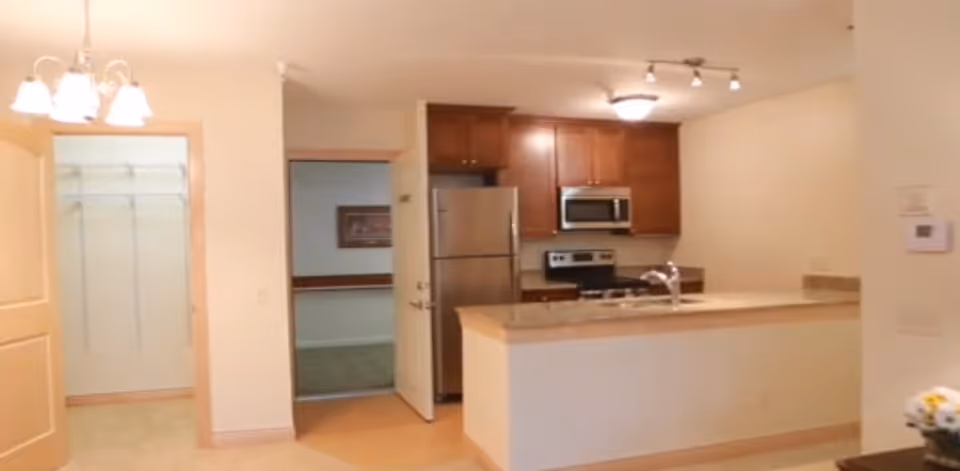 Interior view of a senior living community apartment kitchen area with wooden cabinets, stainless steel refrigerator, microwave, stove, and a countertop with a sink. To the left, there is an open door leading to a walk-in closet with shelving. The walls are painted white and the floor is light-colored.