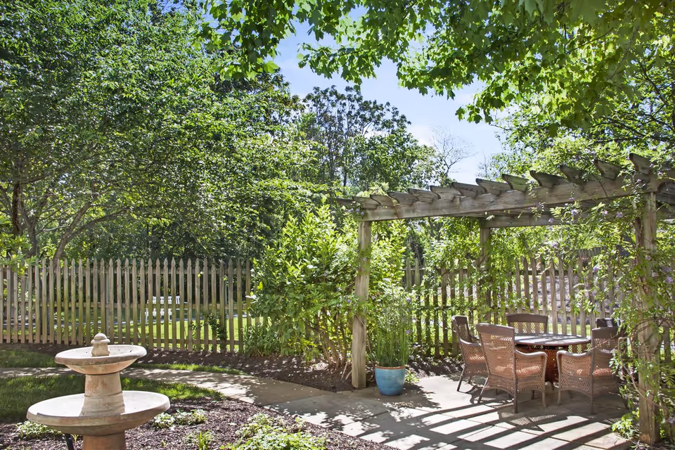Sunlit outdoor garden patio with a wooden pergola, wicker dining set, stone fountain and a picket fence surrounded by trees.