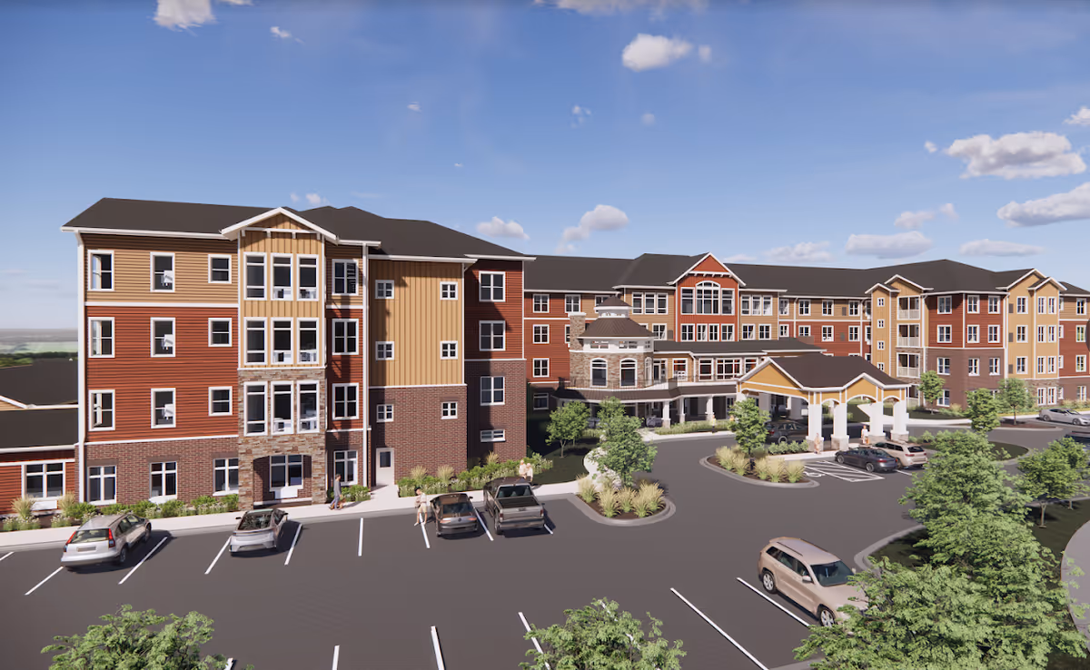 Exterior view of a large, multi-story senior living facility with a mix of brick and siding in earth tones. The building features many windows, a covered entrance with columns, and a parking lot with several cars and a few people walking nearby. The sky is clear with some clouds.