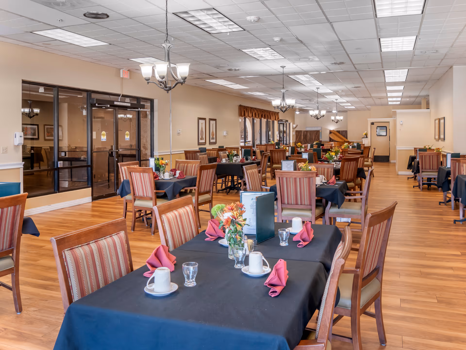 A spacious dining room with multiple tables covered in black tablecloths, each set with cups, glasses, folded red napkins, and flower centerpieces. The room has wooden flooring, striped upholstered chairs, chandeliers hanging from the ceiling, and large windows along one wall.