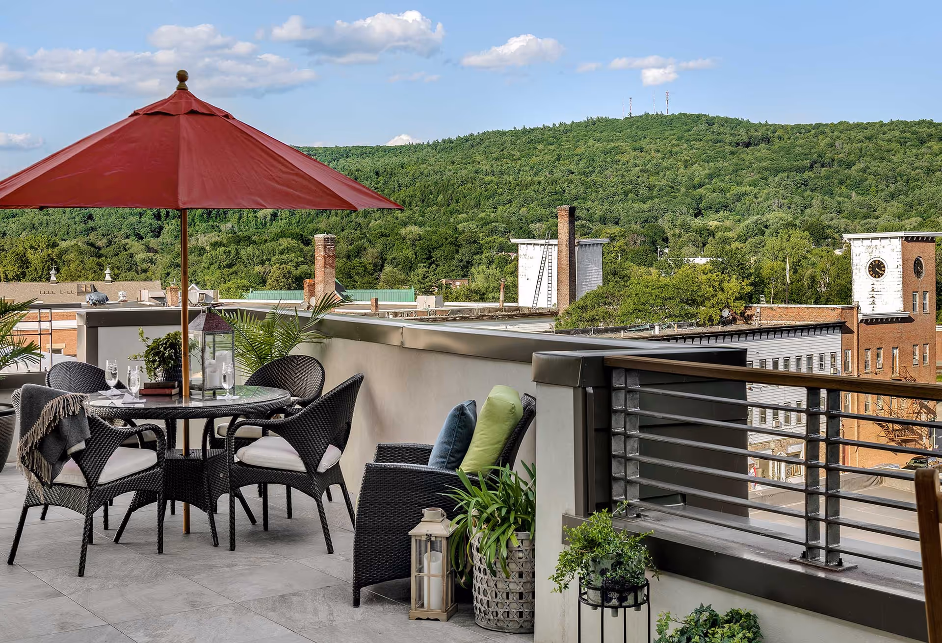 Rooftop patio with a table, wicker chairs and a red umbrella overlooking a small town and tree-covered hills.