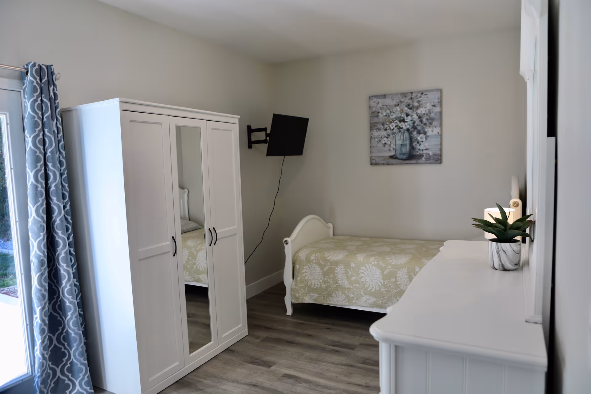 A small, neatly arranged bedroom with a single bed covered in a light green floral bedspread. There is a white wardrobe with a mirrored door, a wall-mounted TV, a white dresser with a small potted plant, and a painting of white flowers on the wall. Blue patterned curtains hang beside a glass door leading outside.