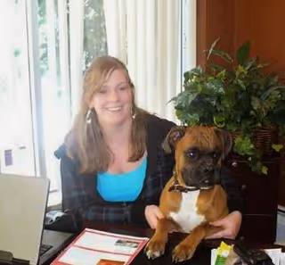 A young woman with light brown hair, wearing a blue top and dark jacket, sitting at a table indoors holding a brown and white dog. There are papers and a laptop on the table, and a large green plant in the background near a window with sheer curtains.
