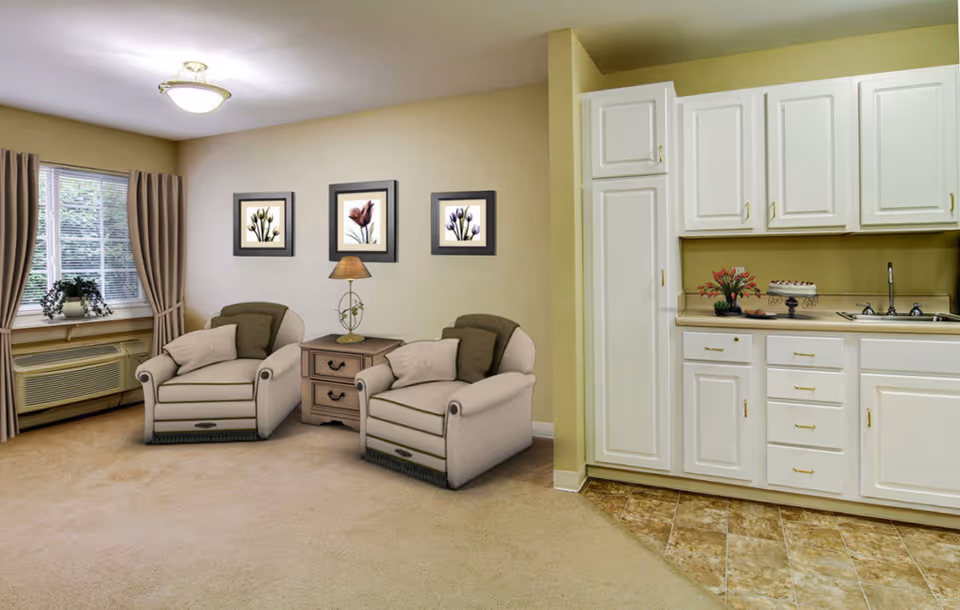 A cozy living area with two beige armchairs separated by a small wooden side table with a lamp. Behind the chairs are three framed floral pictures on a beige wall. To the right is a kitchenette with white cabinets, a sink, and a countertop decorated with a flower vase and a cake. A window with beige curtains and a potted plant is on the left side of the room.